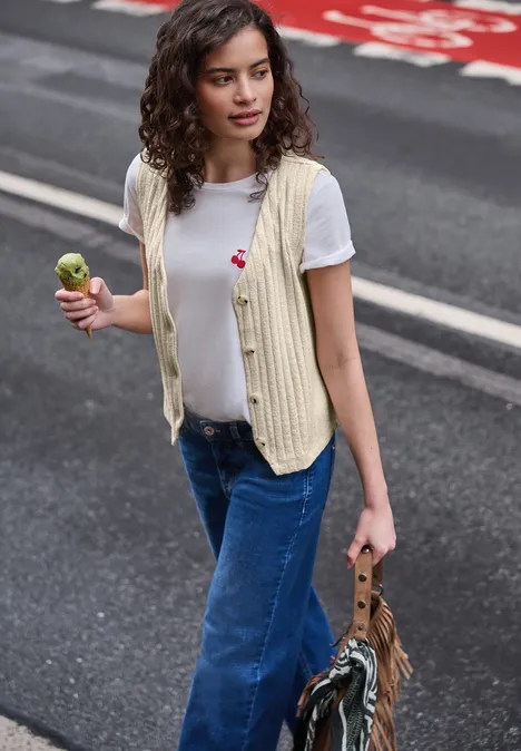 Person in weißem T-Shirt, gestreifter Weste und Jeans läuft über Straße, hält Eis und Tasche.